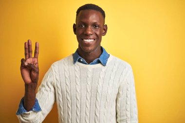 Afro-amerikan adam denim gömlek ve izole sarı arka plan üzerinde beyaz kazak giyen gösteren ve emin ve mutlu gülümserken parmakları ile üç numara işaret.