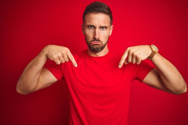 Genç yakışıklı adam kırmızı izole arka plan üzerinde rahat t-shirt giyen üzgün ve üzgün görünümlü aşağı işaret, parmakları ile yön gösteren, mutsuz ve depresif.