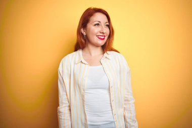 İzole sarı arka plan üzerinde duran çizgili gömlek giyen güzel kızıl saçlı kadın yüzünde gülümseme, doğal ifade ile yan bakıyor. Gülmekten emin.