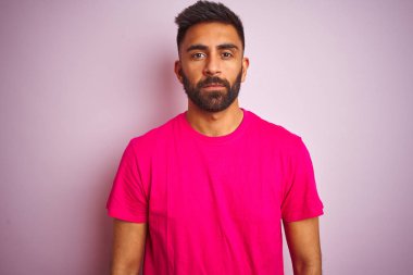 İzole pembe arka plan üzerinde duran t-shirt giyen genç hintli adam yüzünde ciddi ifade ile rahat. Kameraya basit ve doğal bir bakış.