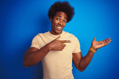 Afro saçlı Amerikalı adam izole mavi arka plan üzerinde çizgili sarı t-shirt giyen hayran ve el ile sunarken kameraya gülümseyerek ve parmak ile işaret.