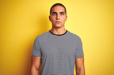 Genç yakışıklı adam sarı izole arka plan üzerinde çizgili t-shirt giyen yüzünde ciddi ifade ile rahat. Kameraya basit ve doğal bir bakış.
