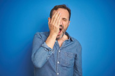 Izole mavi arka plan üzerinde gri saçlı Yakışıklı orta yaş yaşlı adam el ile yarım yüz, göz ve ağız kapsayan yorgun Esneme. Yüz acı içinde acıyor.