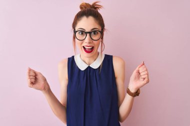Redhead kadın pigtail zarif elbise ve gözlük giyen izole pembe arka plan üzerinde sürpriz kutluyor ve kollarını kaldırdı ve açık gözleri ile başarı için şaşırttı. Kazanan kavramı.