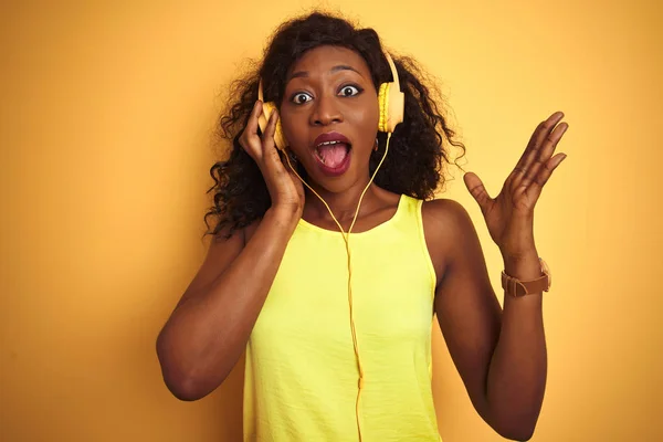 African american woman listening to music using headphones over isolated yellow background very happy and excited, winner expression celebrating victory screaming with big smile and raised hands