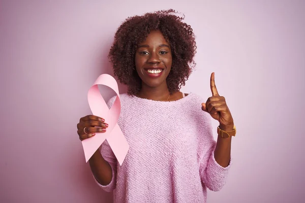 Young african afro woman holding cancer ribbon standing over isolated pink background surprised with an idea or question pointing finger with happy face, number one