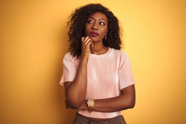 アフリカ系アメリカ人女性は 孤立した黄色の背景の上に立つピンクのTシャツを着て あごに質問を考え ペンシブな表情をしています 思慮深い顔で微笑む 疑いの概念 — ストック写真