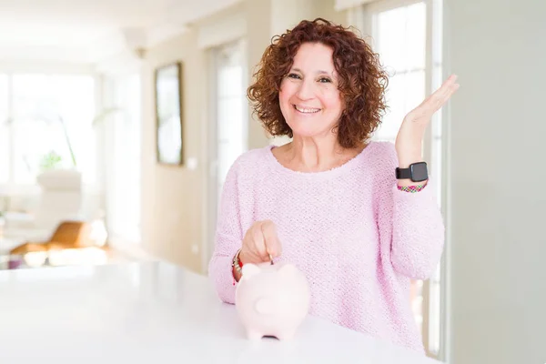 Senior woman saving money putting a coin inside piggy bank very happy and excited, winner expression celebrating victory screaming with big smile and raised hands