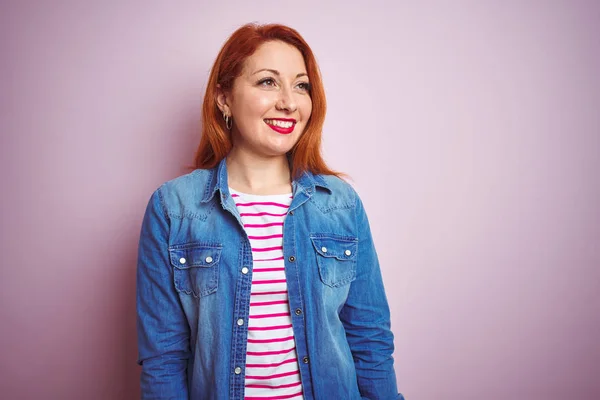 Beautiful redhead woman wearing denim shirt and striped t-shirt over isolated pink background looking away to side with smile on face, natural expression. Laughing confident.