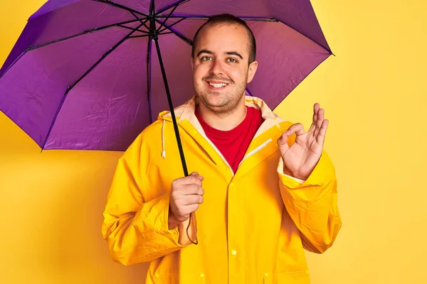 Young man wearing raincoat holding purple umbrella standing over isolated yellow background doing ok sign with fingers, excellent symbol
