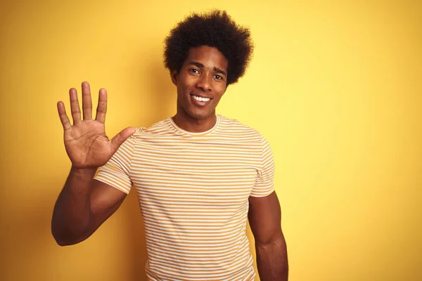 孤立した黄色の背景の上に立つストライプのTシャツを着たアメリカ人男性は 自信を持って幸せに微笑みながら 指番号5を示し 指を上げている — ストック写真
