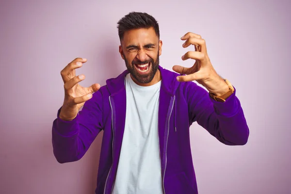 Young indian man wearing purple sweatshirt standing over isolated pink background Shouting frustrated with rage, hands trying to strangle, yelling mad