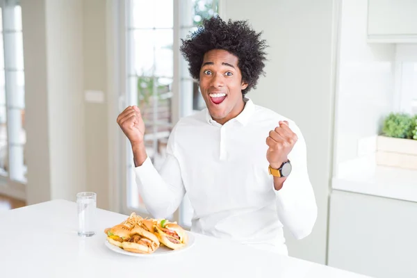 African American hungry man eating hamburger for lunch celebrating surprised and amazed for success with arms raised and open eyes. Winner concept.