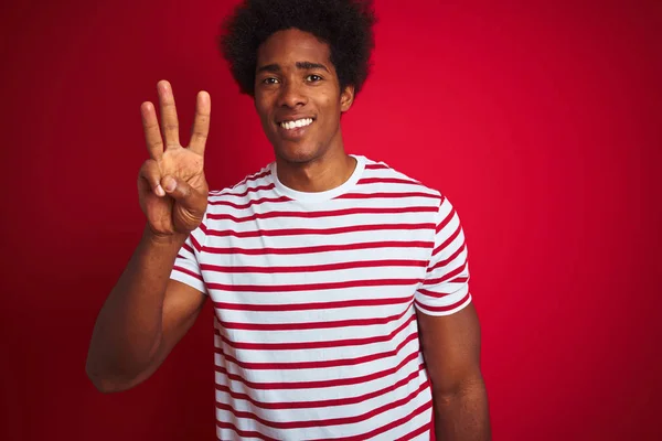 Young african american man with afro hair wearing striped t-shirt over isolated red background showing and pointing up with fingers number three while smiling confident and happy.