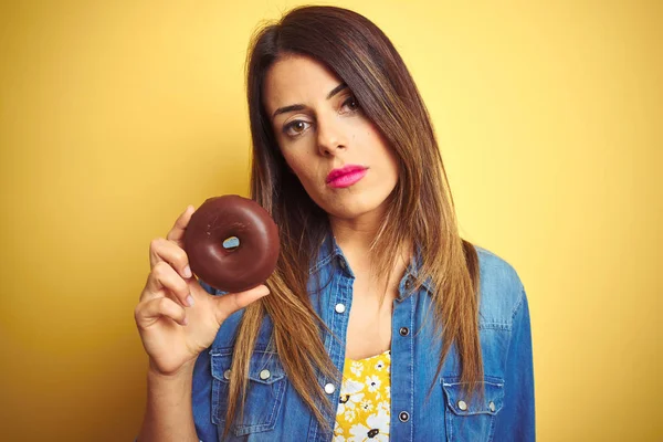Young beautiful woman eating chocolate donut over yellow background with a confident expression on smart face thinking serious