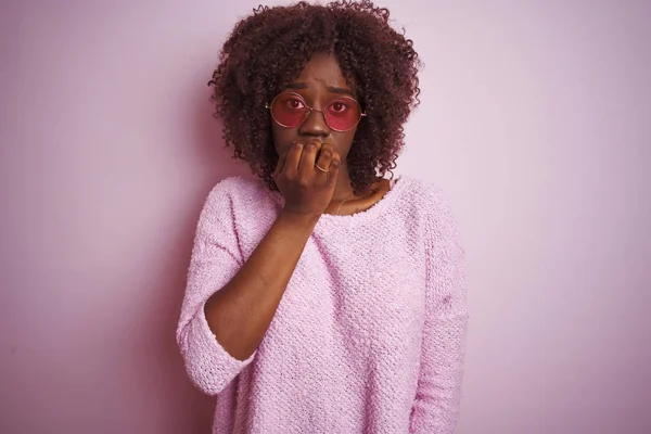 Young african afro woman wearing sweater and sunglasses over isolated pink background looking stressed and nervous with hands on mouth biting nails. Anxiety problem.