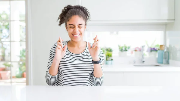 Beautiful african american woman with afro hair wearing casual striped sweater gesturing finger crossed smiling with hope and eyes closed. Luck and superstitious concept.