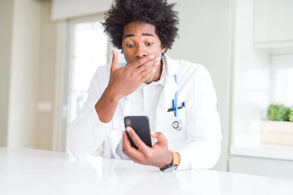 African American doctor man using smartphone at the clinic cover mouth with hand shocked with shame for mistake, expression of fear, scared in silence, secret concept