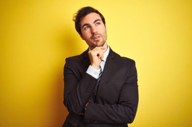 Takım elbise giyen ve kravat takan genç işadamı, elini çenesi üzerinde olan izole sarı arka plan üzerinde durarak soru, dalgın ifade. Düşünceli bir yüzle gülümsüyor. Şüphe kavramı.
