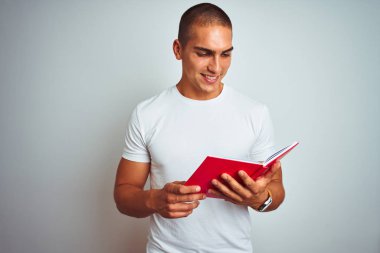 Genç yakışıklı adam ayakta mutlu bir yüz ile beyaz izole arka plan üzerinde kırmızı kitap okuma ve dişleri gösteren kendinden emin bir gülümseme ile gülümseyerek