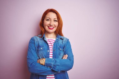 Denim gömlek giyen güzel kızıl saçlı kadın ve izole pembe arka plan üzerinde çizgili t-shirt mutlu yüz kameraya bakarak çapraz kolları ile gülümseyerek. Pozitif kişi.