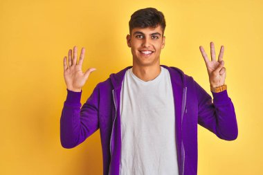 Mor sweatshirt giyen genç hintli adam izole sarı arka plan üzerinde duran gösteren ve parmakları ile işaret ederken kendinden emin ve mutlu gülümseyerek sekiz.