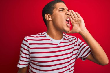 Genç yakışıklı Arap adam izole kırmızı arka plan üzerinde çizgili t-shirt giyen bağırarak ve ağız da el ile yan yüksek sesle bağırıyor. İletişim kavramı.