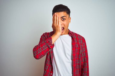 Bir gözünü eliyle kaplayan izole beyaz arka plan üzerinde duran kırmızı gömlek giyen genç Brezilyalı adam, yüzünde kendine güvenen bir gülümseme ve sürpriz duygu.