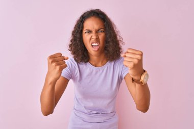 Genç Brezilyalı kadın izole pembe arka plan üzerinde duran t-shirt giyen kızgın ve öfkeli öfkeli yumruklar yükselterek öfke ile bağırırken. Öfke ve agresif kavram.