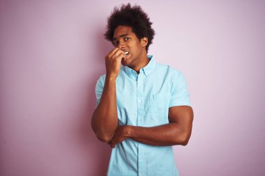 Afro saçlı, mavi gömlek giyen genç amerikalı adam izole pembe arka plan üzerinde duran stresli ve ağız tırnak ısırma elleri ile sinirli görünümlü. Anksiyete sorunu.