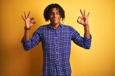 Izole sarı arka plan üzerinde duran rahat gömlek giyen dreadlocks ile Afro adam rahat ve gözleri parmakları ile meditasyon jest yapıyor kapalı gülümseyerek. Yoga konsepti.
