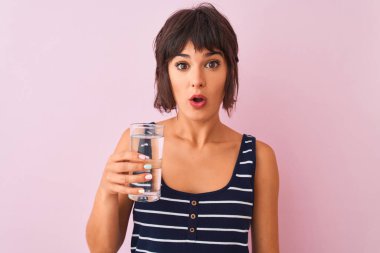 İzole edilmiş pembe arka plan üzerinde duran su bardağı tutan genç güzel kadın sürpriz bir yüzle şoktan korkmuş, korku ve korku yla heyecanlı