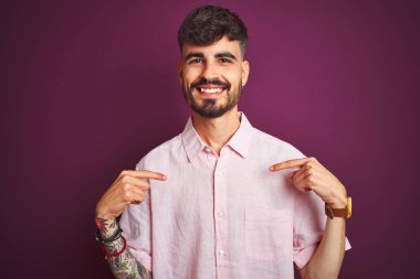 Yazlık pembe gömlek giyen dövmegiyen genç adam izole mor arka plan üzerinde duran yüzünde gülümseme ile emin görünümlü, parmakları gururlu ve mutlu kendini işaret.