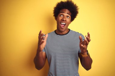 Afro saçlı Afro-Amerikalı adam izole sarı arka plan üzerinde lacivert çizgili t-shirt giyen deli ve deli bağırarak ve agresif ifade ve kollar kaldırdı ile bağırıyor. Hayal kırıklığı kavramı.