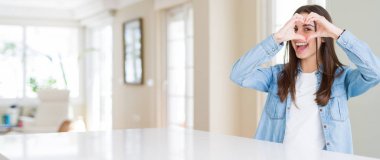 Evde beyaz masada oturan güzel genç kadının geniş açılı resmi El ve parmaklarla kalp şekli yapmak burcuna bakarak gülümseyerek