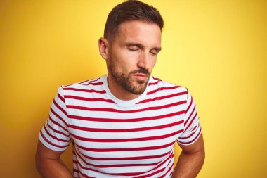 Genç yakışıklı adam mide üzerinde el ile sarı izole arka plan üzerinde rahat kırmızı çizgili t-shirt giyiyor çünkü mide, ağrılı hastalık kötü hissediyorum. Ağrı kavramı.