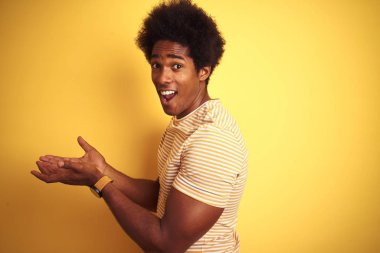 Afro saçlı afro saçlı adam izole sarı arka plan üzerinde duran çizgili t-shirt giyen elleri açık avuç içi ile bir yana fotokopi alanı gösteren, reklam gülümseyerek sunan mutlu mutlu