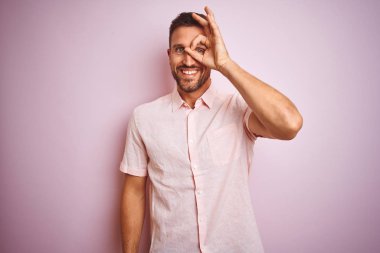 Genç yakışıklı adam pembe izole arka plan üzerinde zarif yazlık gömlek giyen el gülümseyerek ok jest yapıyor, mutlu yüz ile parmaklardan bakan göz.
