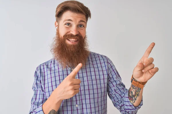 Redhead İrlandalı adam izole beyaz arka plan üzerinde kablosuz kulaklık kullanarak müzik dinlerken gülümseyerek ve yan iki el ve parmak ile işaret kamera bakıyor.