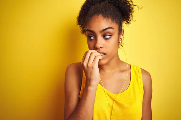 African american woman listening to music using earphones over isolated yellow background looking stressed and nervous with hands on mouth biting nails. Anxiety problem.