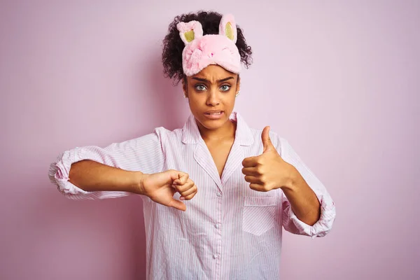 年轻的非洲裔美国妇女穿着睡衣和面具在孤立的粉红色背景做大拇指上下 分歧和协议表达 疯狂的冲突 — 图库照片
