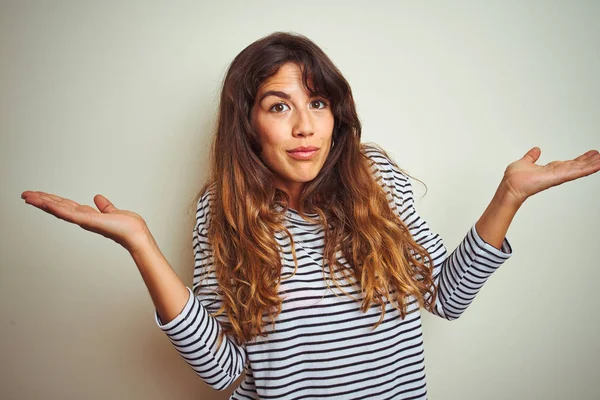 Genç güzel kadın beyaz izole arka plan üzerinde çizgili t-shirt giyen habersiz ve kollar ve eller kaldırdı ile karışık ifade. Şüphe kavramı.