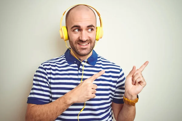 孤立した背景の上に黄色のヘッドフォンを着用して音楽を聴いている若者は微笑み 両手と指で側面を指差すカメラを見ている — ストック写真