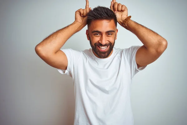 Young indian man wearing t-shirt standing over isolated white background doing funny gesture with finger over head as bull horns