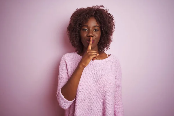 Young african afro woman wearing sweater standing over isolated pink background asking to be quiet with finger on lips. Silence and secret concept.