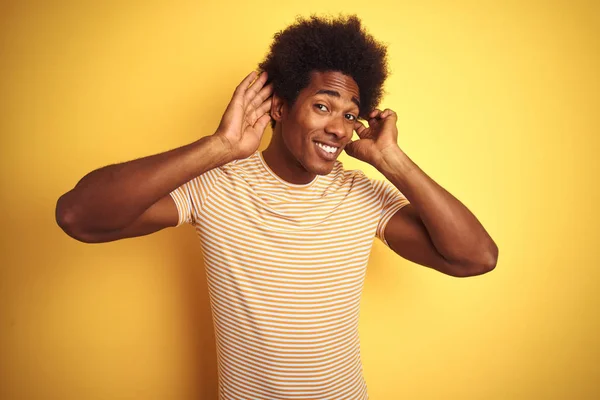 孤立した黄色の背景の上に立つストライプのTシャツを着たアメリカ人男性は 耳のジェスチャーで両手を聞こうとし ゴシップに興味を持っています 聴覚障害 聴覚障害 — ストック写真