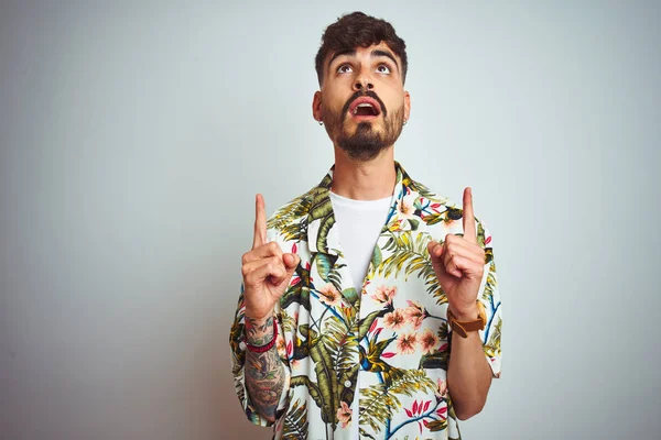 Tatilde yazlık gömlek giyen dövmeli adam izole beyaz arka plan üzerinde duran şaşırmış ve şaşırmış yukarı bakarak ve parmakları ile işaret ve kolları kaldırdı.