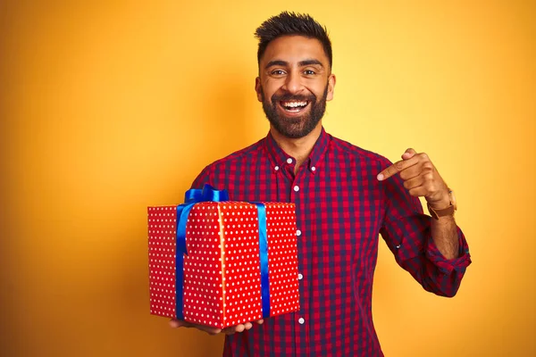 Arab indian hispanic man holding birthday gift standing over isolated yellow background with surprise face pointing finger to himself