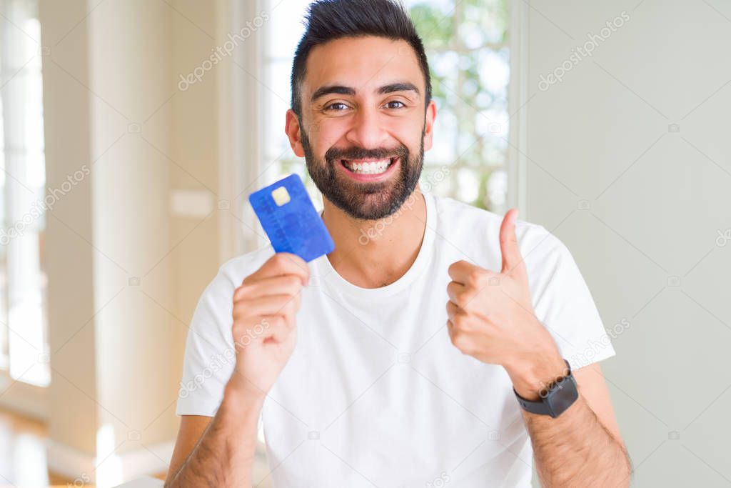 Handsome hispanic man holding credit card happy with big smile doing ok sign, thumb up with fingers, excellent sign