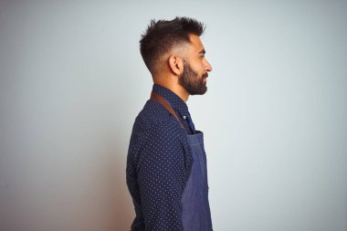 Yan bakarak izole beyaz arka plan üzerinde duran önlük üniforma giyen genç hintli çalışan adam, kendine güvenen bir gülümseme ile doğal yüzü ile profil poz dinlenmek.